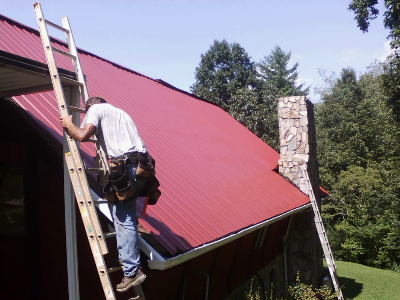 Roofer on a ladder beside a red metal roof and stone chimney — Soffit & Fascia Repair by Roofing Brothers in Ten Sleep